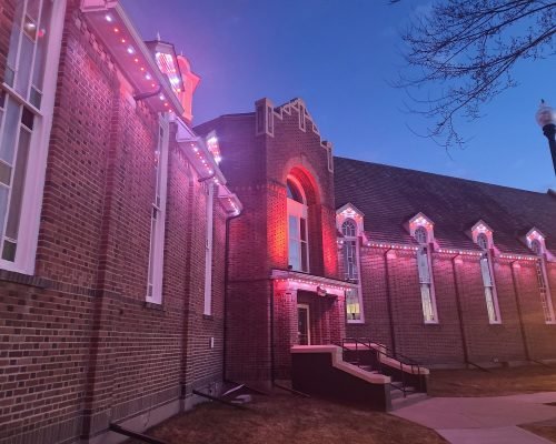 Vibrant pink LED architectural lighting illuminates historic red brick church with arched windows and gabled roofline at twilight, custom installation by Spectrum Lighting Inc.