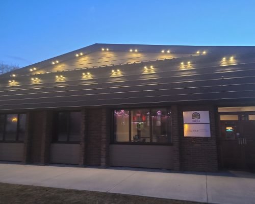 Evening view of Spectrum Lighting Inc. commercial building at dusk, beautifully illuminated with warm white LED string lights along the roofline and eaves, brick facade, large glowing windows, and entrance sign – expert lighting installation by spectrumlightinginc.com