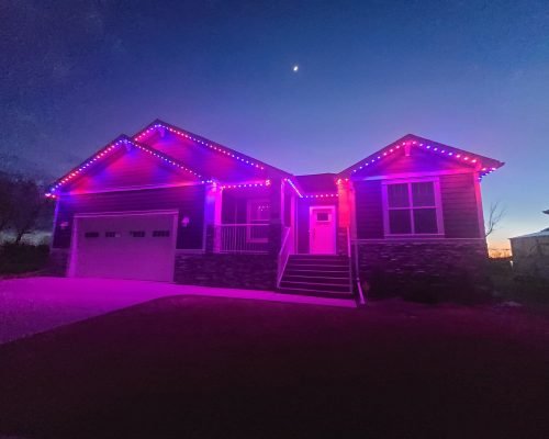 Vibrant purple and pink LED holiday lights illuminating a modern stone-accented house at night, outlining roofline, garage doors, windows, and entry steps. Professional installation by Spectrum Lighting Inc., with crescent moon in twilight sky.