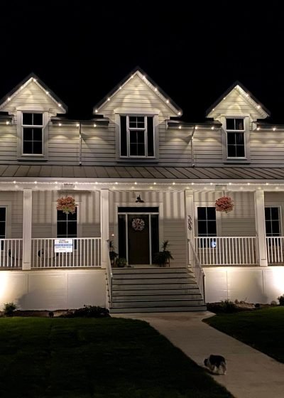 Stunning nighttime view of a white farmhouse-style home with professional Spectrum Lighting Inc. white LED string lights outlining gabled roofs, wraparound porch, railings, and front steps. Features hanging floral baskets, Christmas wreath on door, uplighting, lush green lawn, pathway, and subtle holiday glow.