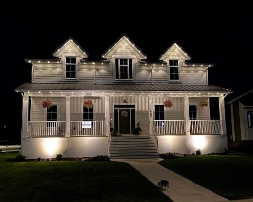 Stunning nighttime view of a white farmhouse-style home with professional Spectrum Lighting Inc. white LED string lights outlining gabled roofs, wraparound porch, railings, and front steps. Features hanging floral baskets, Christmas wreath on door, uplighting, lush green lawn, pathway, and subtle holiday glow.