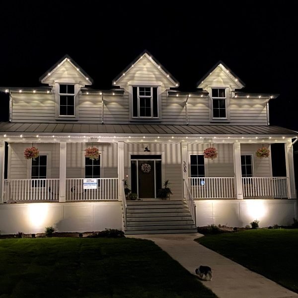 Stunning nighttime view of a white farmhouse-style home with professional Spectrum Lighting Inc. white LED string lights outlining gabled roofs, wraparound porch, railings, and front steps. Features hanging floral baskets, Christmas wreath on door, uplighting, lush green lawn, pathway, and subtle holiday glow.
