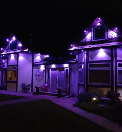 Vibrant purple LED exterior lighting on a modern gabled house at night by Spectrum Lighting Inc., illuminating rooflines, walls, patio furniture, pathway, and landscaped garden for stunning nighttime glow