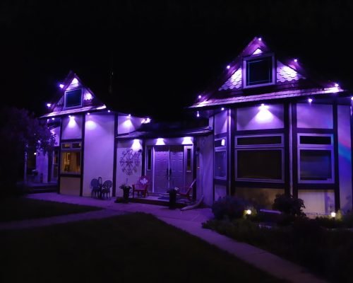 Vibrant purple LED exterior lighting on a modern gabled house at night by Spectrum Lighting Inc., illuminating rooflines, walls, patio furniture, pathway, and landscaped garden for stunning nighttime glow