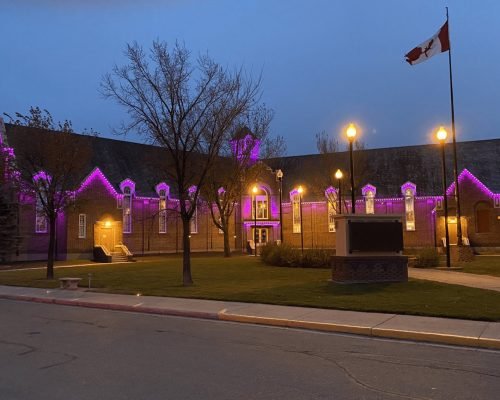 Vibrant purple LED architectural lighting illuminates a historic gabled building at dusk, with glowing rooflines and windows, Canadian flag, trees, street lamps, and monument by Spectrum Lighting Inc.