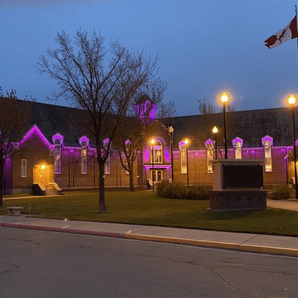 Vibrant purple LED architectural lighting illuminates a historic gabled building at dusk, with glowing rooflines and windows, Canadian flag, trees, street lamps, and monument by Spectrum Lighting Inc.