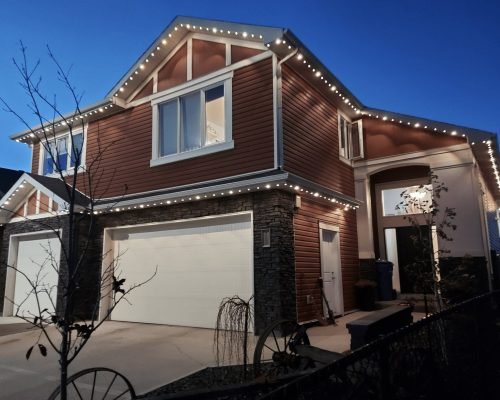 Elegant two-story red-sided home with stone accents and two-car garage, glowing with professional white LED string lights along rooflines and eaves at dusk, holiday lighting installation by Spectrum Lighting Inc.
