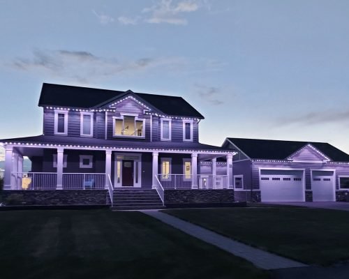 Elegant two-story home with wrap-around porch, attached garage, and lush lawn illuminated by vibrant purple LED accent lighting at dusk – professional outdoor landscape lighting by Spectrum Lighting Inc.