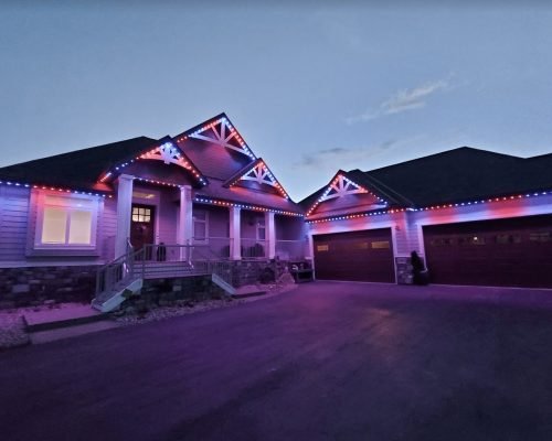 Patriotic red, white, and blue LED lighting illuminating a modern two-story home at dusk. Vibrant lights outline gabled rooflines, stone accents, front porch columns, large windows, and double garage doors on a driveway. Custom holiday lighting display by Spectrum Lighting Inc.