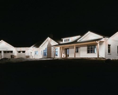 Stunning nighttime view of modern white farmhouse home illuminated by professional exterior lighting from Spectrum Lighting Inc. Features three-car garage, wrap-around porch, large windows, beautifully lit against dark sky for custom outdoor lighting design.