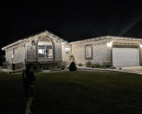 Nighttime view of a beautifully lit stucco home with soft white LED landscape lighting outlining rooflines, arched windows, garage, and eaves. Professional exterior illumination by Spectrum Lighting Inc. enhances modern architecture, front yard shrubs, and grass.
