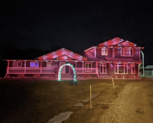 Vibrant pink holiday lights illuminating a large ranch-style house at night, featuring white roofline outlines, green arched entrance garland, and professional Christmas lighting installation by Spectrum Lighting Inc.