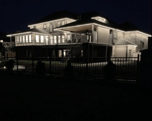 Luxurious elevated waterfront home at night, illuminated with custom white LED architectural lighting outlining rooflines, eaves, decks, garage doors, sunroom, balcony, and black iron fence by Spectrum Lighting Inc.