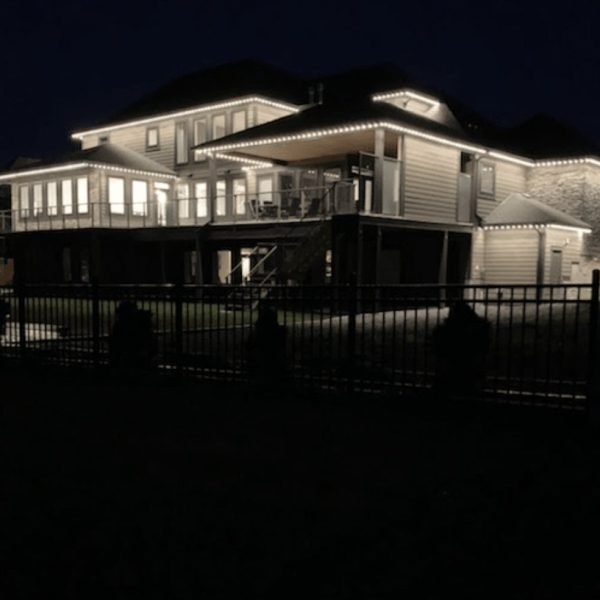 Luxurious elevated waterfront home at night, illuminated with custom white LED architectural lighting outlining rooflines, eaves, decks, garage doors, sunroom, balcony, and black iron fence by Spectrum Lighting Inc.