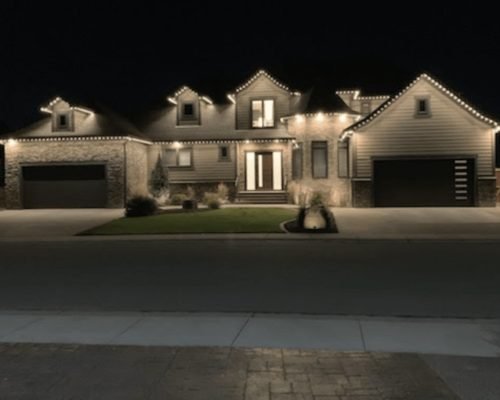 Stunning nighttime view of a modern two-story home with stone and siding facade, beautifully illuminated by warm white LED landscape lighting along rooflines, eaves, windows, and garage doors from Spectrum Lighting Inc.
